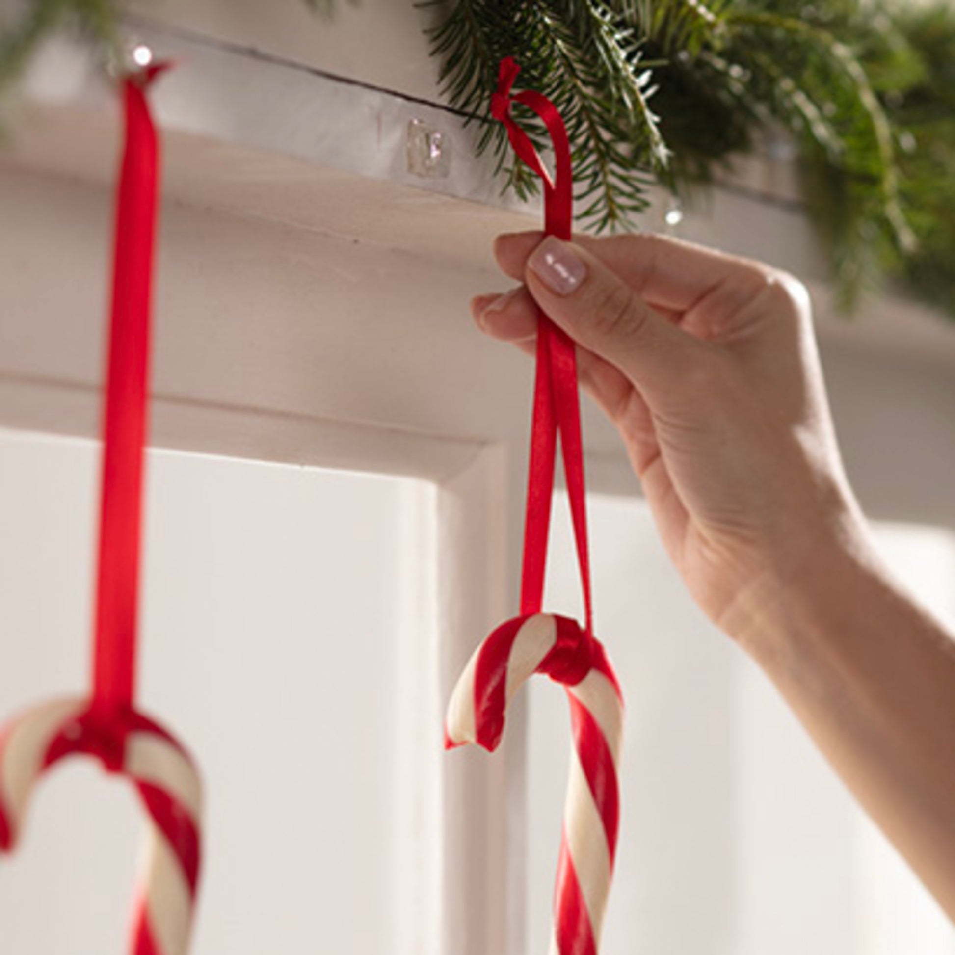 Eine Hand hängt eine rot-weiße Zuckerstange an einem roten Band auf einer weißen Fläche auf, mit Command™ Mini-Haken, transparent, mit transparenten Strips, 17006CLRFGN von 3M Deutschland GmbH. Ein weiterer Zuckerstab hängt bereits daneben an grünem Tannengirlande.