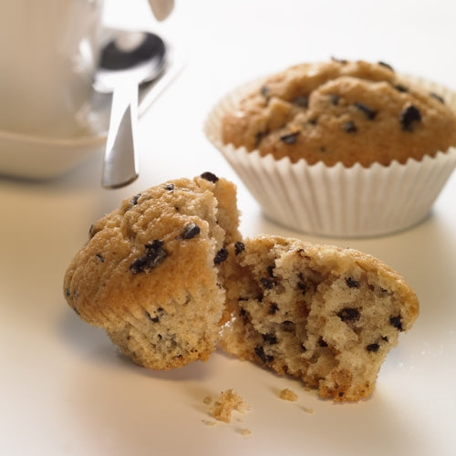 Op de voorgrond is een gehalveerde Chocolate Chip Muffin te zien, waarover kruimels verspreid zijn. Op de achtergrond ligt een andere muffin in een PAPSTAR 1000 bakvormpje rond van PAPSTAR GmbH, naast een witte kop met schotel en lepel op een witte ondergrond.
