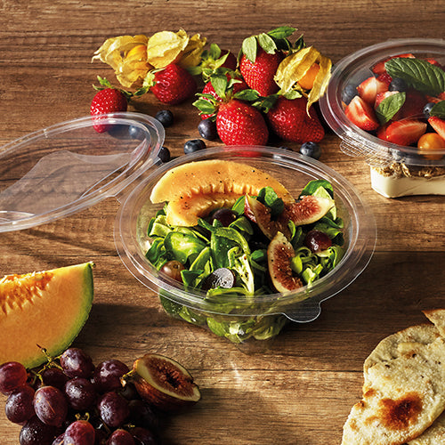 Op een houten tafel staat een ronde Starpak 50 delicatessen- en slaschalen met klapdeksel (R-PET glashelder) met salade van meloen, vijgen en bladgroenten. Daarnaast staan aardbeien, druiven, meloenstukjes, cherrytomaten, platbrood en een andere schaal met tomaten en basilicum.