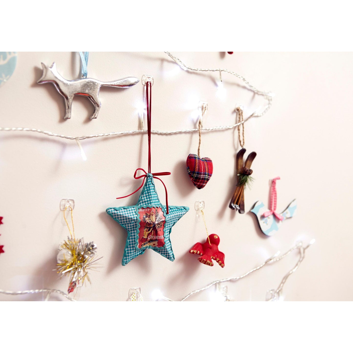 Aan een beige muur hangen witte lichtsnoeren en feestelijke ornamenten - waaronder een zilveren vos, een blauwe ster, een geruit hart en een witte duif - aan transparante Command™ decoratieclips van 3M Deutschland GmbH.