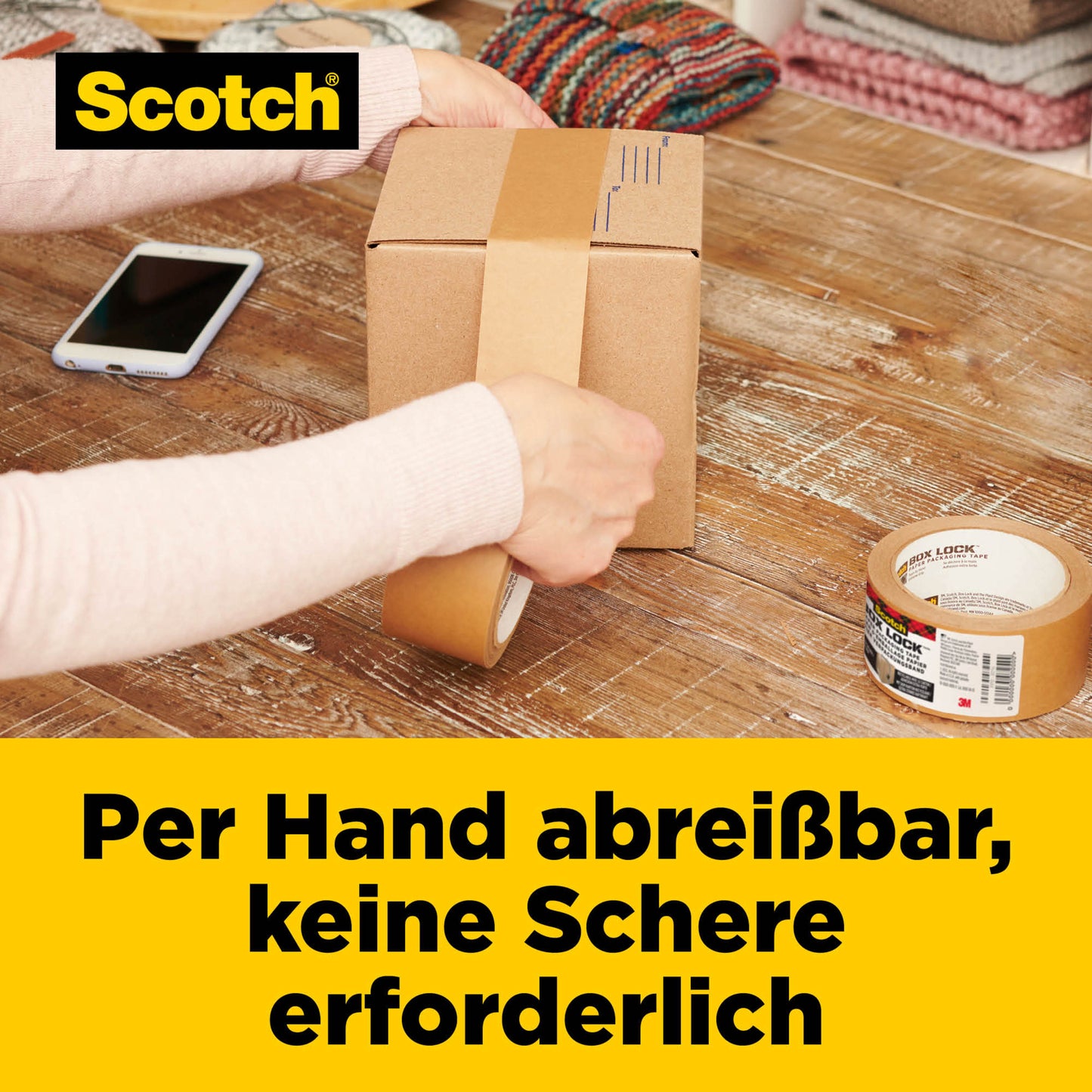 Eine Person reißt Scotch® Box Lock™ Papier-Verpackungsband mit der Hand ab, um eine Kiste auf einem Holztisch zu verschließen. Zu sehen sind eine Rolle Klebeband, ein Smartphone und gefaltete Textilien. Der deutsche Text hebt die Easy-Tear-Funktion der 3M Deutschland GmbH hervor.
