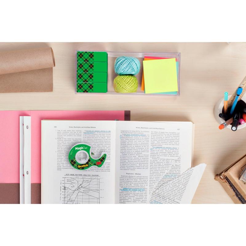 Een bureau met een geopend boek, Scotch® Magic™ Onzichtbare Plakband Voordeelpak (24 rollen, 3M Deutschland GmbH), gekleurd papier, notitieblaadjes, touw, gum, een beker met pennen en potloden, evenals gestapelde boeken en gerold bruin papier.