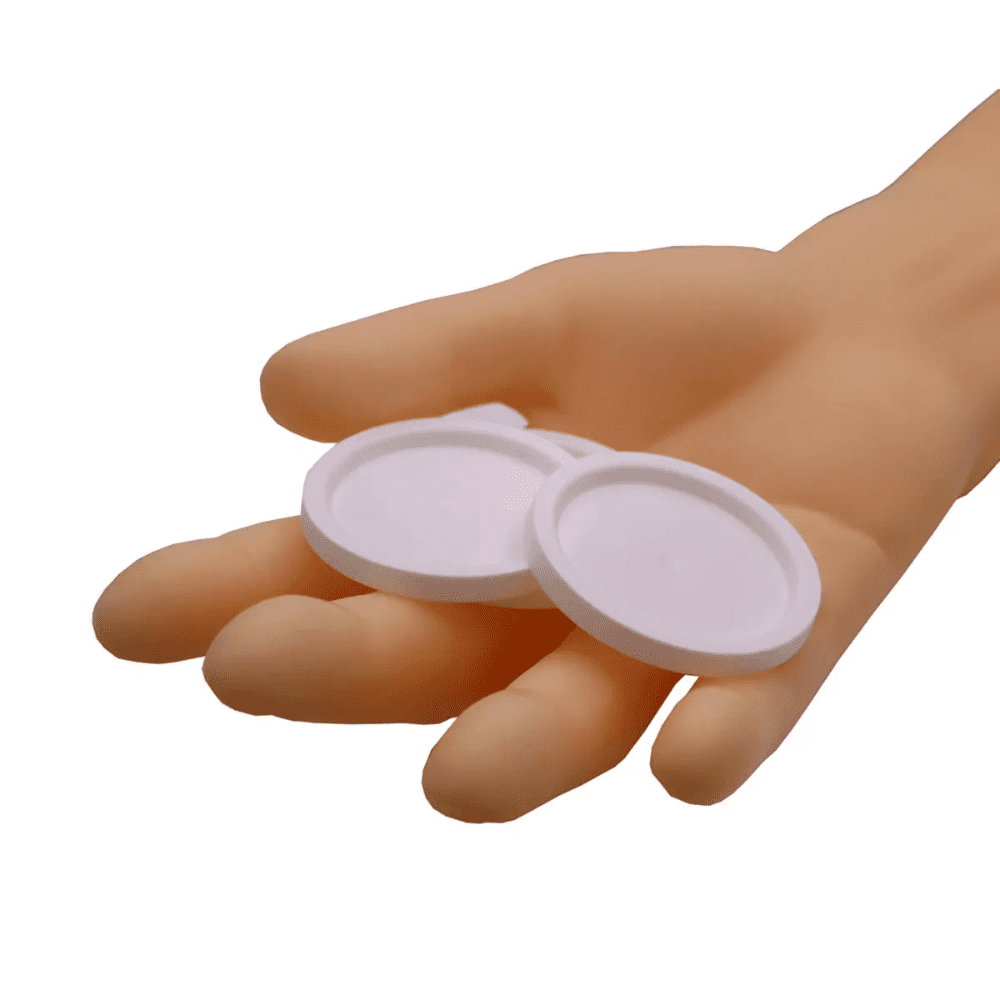Een hand houdt twee ronde AMPri deksels voor medicijnbekers, witte objecten tegen een eenvoudige witte achtergrond.