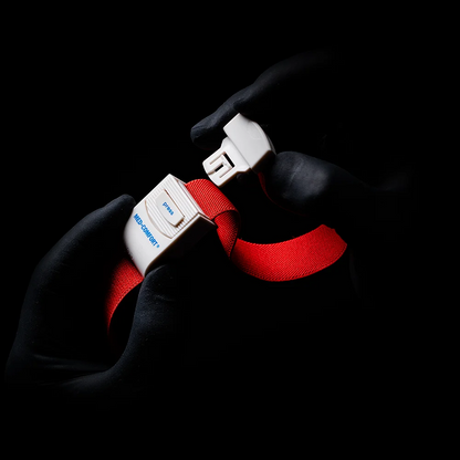 Met handschoenen bedekte handen, die aan een AMPri MED-COMFORT tourniquet doen denken, grijpen voor een schaduwrijke achtergrond de gesp van een felrode veiligheidsgordel. Op de gesp zijn het woord 'Blue' en de letter 'B' te zien.