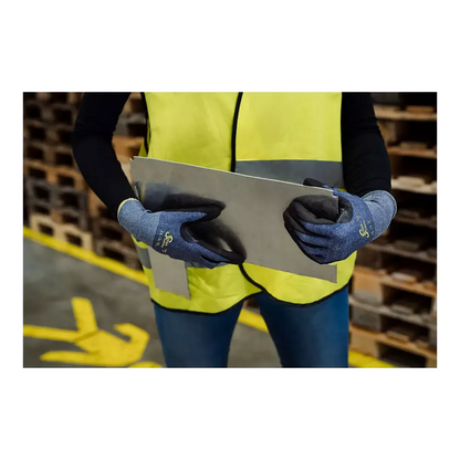 Een persoon met een neongeel veiligheidsvest en blauwe AMPri SolidSafety Cut snijbestendige handschoenen S - XXL van AMPri Handelsgesellschaft mbH hanteert voorzichtig een grote metalen plaat in het magazijn. Op de achtergrond zijn houten pallets gestapeld, gele markeringen op de vloer leiden veilige bewegingen en benadrukken de arbeidsveiligheid.
