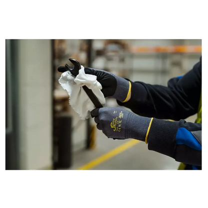 Een persoon met AMPri SolidSafety Tough Air ultralichte montagehandschoenen van AMPri Handelsgesellschaft mbH reinigt binnenshuis voorzichtig een moersleutel met een witte doek. De grijze handschoenen, die bekend staan om hun slijtvastheidsniveau 4, steken af tegen de wazige industriële rekken en apparaten op de achtergrond.