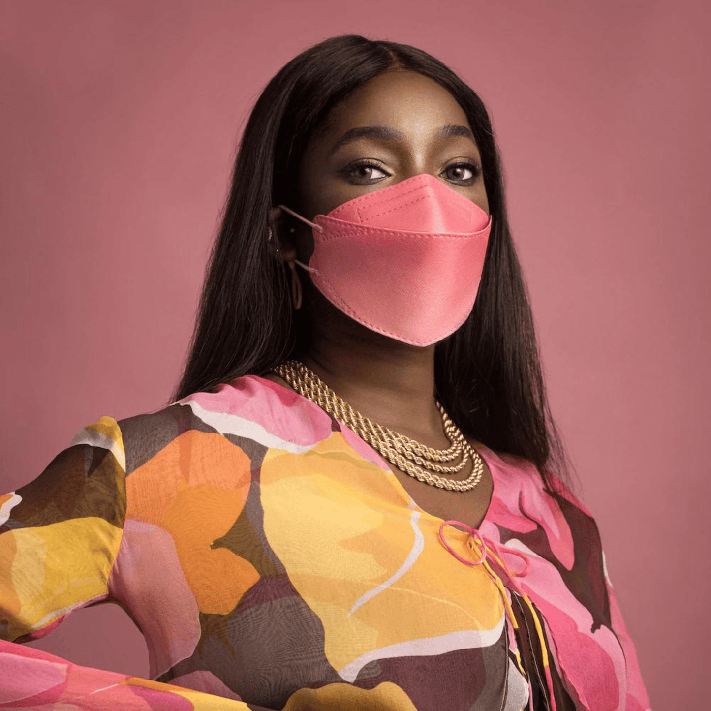 Een persoon draagt een deeltjesfilterende Altruan Designer-gezichtsmasker (FFP2-norm) in een stijlvolle roze tint en een kleurrijke top met abstract patroon voor een roze achtergrond. Ze heeft lang, donker haar en draagt meerdere gouden kettingen.