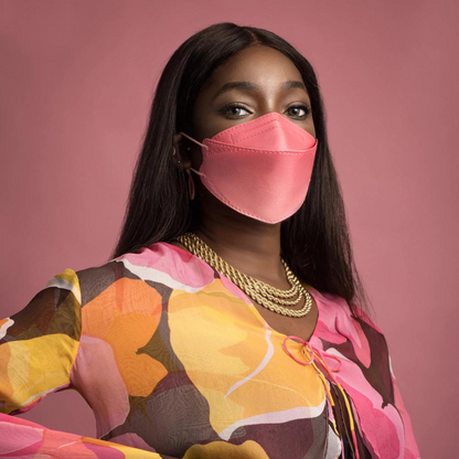 Een persoon draagt een deeltjesfilterende Altruan Designer-gezichtsmasker (FFP2-norm) in een stijlvolle roze tint en een kleurrijke top met abstract patroon voor een roze achtergrond. Ze heeft lang, donker haar en draagt meerdere gouden kettingen.