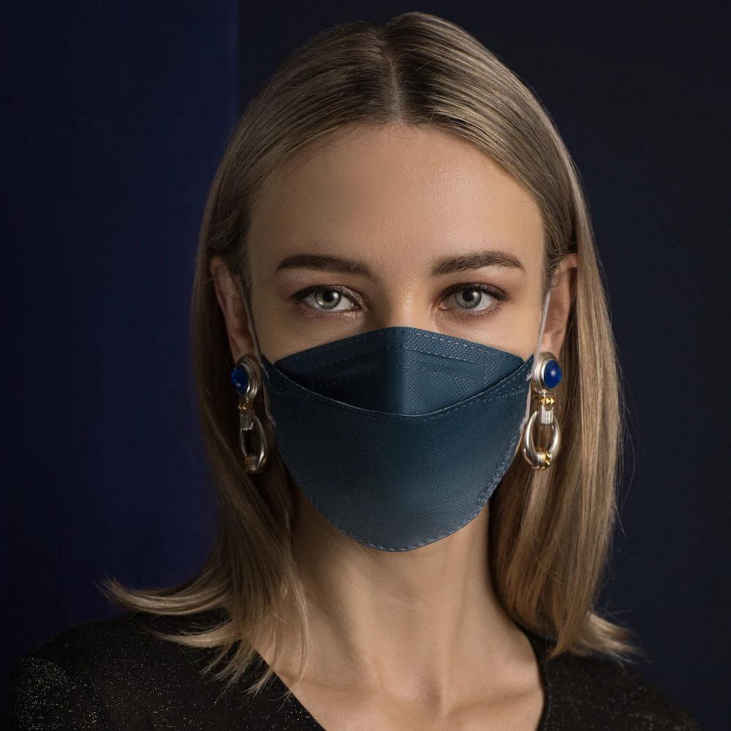 Een persoon met lang haar draagt een Altruan Designer-gezichtsmasker Deeltjesfilterend in blauw, die effectieve bescherming biedt volgens de FFP2-norm. Ze combineert het met grote decoratieve oorbellen voor een donkere achtergrond en draagt een zwart shirt.