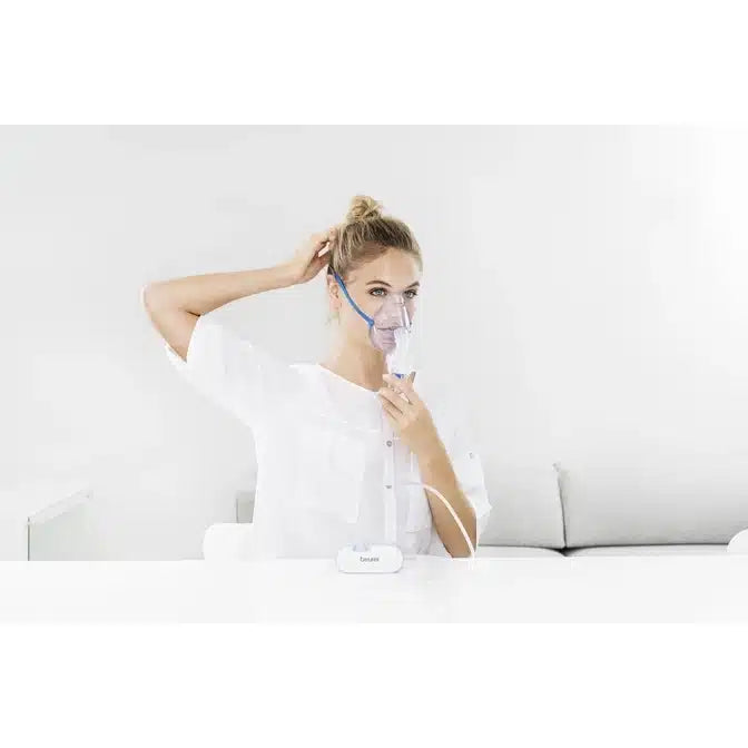 Een persoon gebruikt het uitloopartikel: Beurer kleine inhalator IH 60 van Beurer GmbH met een gezichtsmasker aan een witte tafel en doet haar haar. De minimale witte achtergrond benadrukt dit compacte apparaat voor ademhalingsaandoeningen.