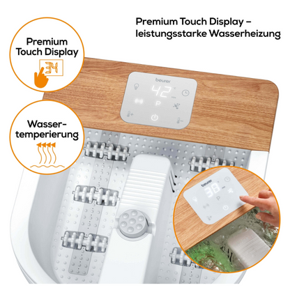 Een voetbad met het merk Beurer FB 65 Wellness voetbubbelbad van Beurer GmbH heeft een witte basis en een houten plaat, met een digitaal touch-display dat een temperatuur van 42 °C aangeeft. Op de etiketten staat 'Watertemperering' en 'Premium Touch Display'. Een hand past het display aan en massagerollen voor vibratie- en bubbelmassage zijn zichtbaar.