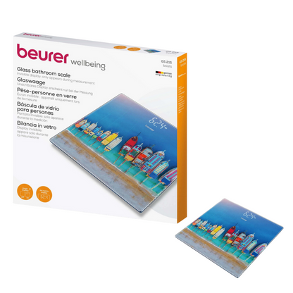 De afbeelding toont de Beurer GS 215 glasweegschaal van Beurer GmbH zowel in de verpakking als afzonderlijke weegschaal. De weegschaal toont een kleurrijke illustratie van strandhuisjes en een vuurtoren, evenals een magisch display voor betere zichtbaarheid. De verpakking met de opdruk "Beurer GS 215 glasweegschaal | Verpakking (1 stuk)" biedt productinformatie in meerdere talen.