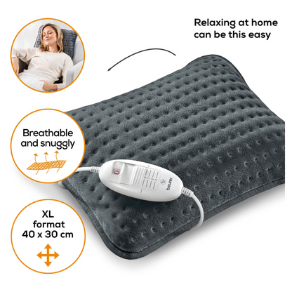 Een vrouw ligt op een bank en glimlacht met gesloten ogen. Naast haar staat een Beurer HK 48 Sofa-verwarmingskussen Cosy in donkergrijs met een witte bedieningsunit. Op het kussen staat 'Zo eenvoudig kan ontspannen thuis zijn', 'Ademend en knus' en 'XL-formaat 40 x 30 cm'.