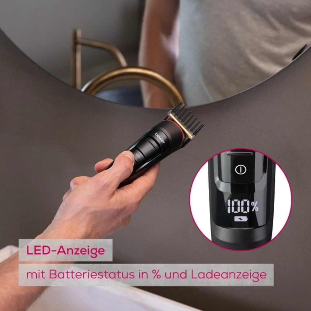 Een hand houdt het uitloopartikel Beurer Multigroomer MN9X van Beurer GmbH met LED-display, dat de accustatus toont, voor de badkamerspiegel. Duitse opschrift wijst op het waterdichte apparaat en de laadstatus.