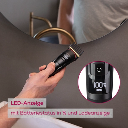 Een hand houdt het uitloopartikel Beurer Multigroomer MN9X van Beurer GmbH met LED-display, dat de accustatus toont, voor de badkamerspiegel. Duitse opschrift wijst op het waterdichte apparaat en de laadstatus.