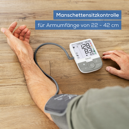 Een gebruiker werkt met het Beurer GmbH uitloopartikel: bovenarm-bloeddrukmeter BM 53 op een lichte houten ondergrond. Het display toont "Manchetpositiecontrole voor armomtrekken van 22-42 cm" en wijst op aritmieherkenning en beroertepreventie.