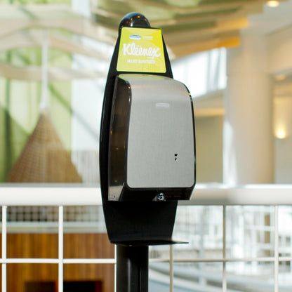 Een Kimberly-Clark GmbH Elektronische Waslotionen Standaard (Zwart, 1 dispenser) is binnen geplaatst nabij een wit metalen hek voor hygiënische handreiniging, met onscherpe architectonische details op de achtergrond.