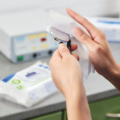 Een persoon reinigt een medisch instrument met Hartmann Bacillol® 30 Sensitive Green Tissues Snelle Desinfectiedoekjes van Paul Hartmann AG. Op de achtergrond zijn een verpakking desinfectiedoekjes en verschillende medische apparaten te zien. De omgeving lijkt een schone, klinische omgeving te zijn, wat het gebruik van milieuvriendelijke, materiaalvriendelijke desinfectiedoekjes benadrukt.