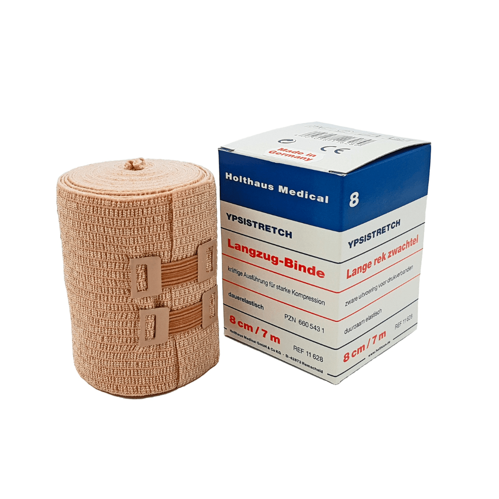 Naast de verpakking is een medische elastische zwachtel afgebeeld. Op de doos staat "Holthaus Ypsistretch Langzug-Binde, sterk" en de gegevens zijn in het Duits. De huidkleurige zwachtel van Holthaus Medical GmbH & Co. KG biedt een effectieve compressie en wordt met twee kunststofsluitingen bevestigd.
