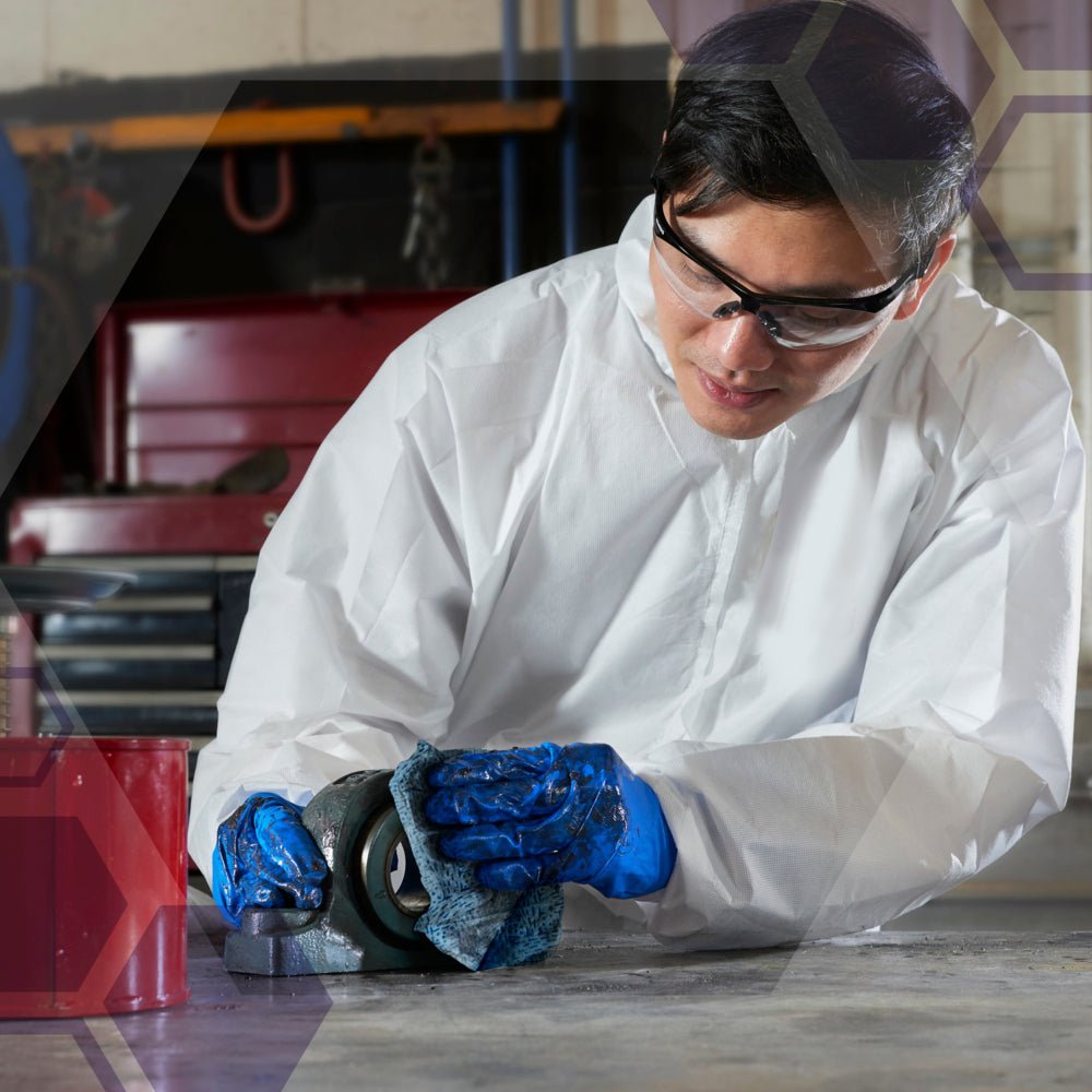 Een persoon met veiligheidsbril, wit pak en blauwe handschoenen knielt op een betonnen vloer in een industriële reinigingsomgeving en gebruikt de Kimtech® procesreinigingsdoeken - BRAG™ Box van Kimberly-Clark GmbH om een metalen object met een doek te reinigen.