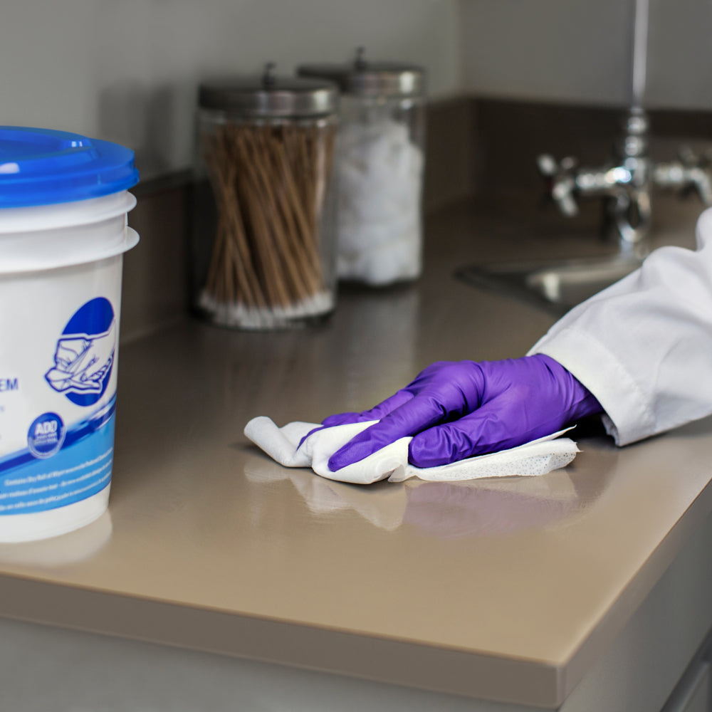 Eine Person mit lila Handschuhen wischt mit WypAll® Wettask™ Wischtüchern der Kimberly-Clark GmbH (6 Rollen x 90 Blatt) eine beigefarbene Arbeitsplatte in der Nähe des Waschbeckens ab, während im Hintergrund Gläser mit Wattestäbchen und -bällen zu sehen sind.