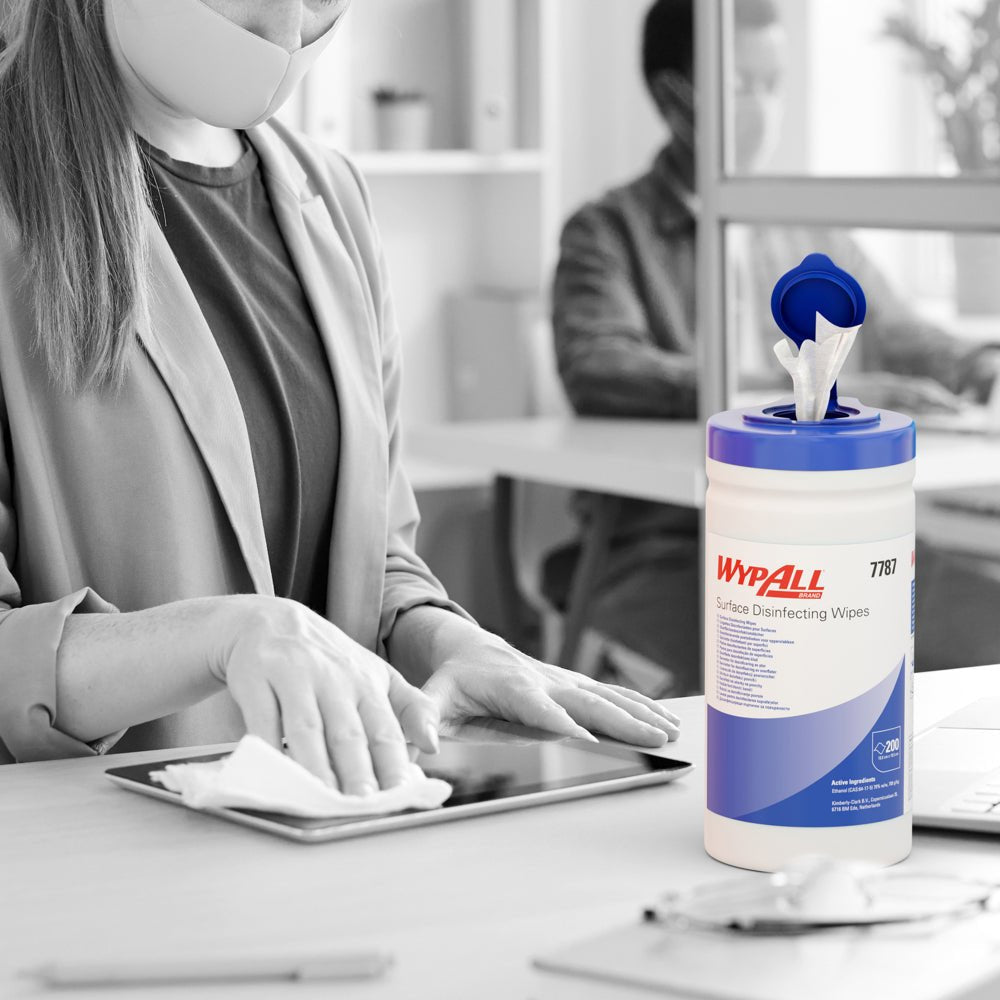 Een gemaskerde persoon desinfecteert met WypAll® oppervlaktedesinfectiedoekjes (dispenseremmer, 200 vellen) van Kimberly-Clark GmbH een tablet op een bureau, terwijl de bak zichtbaar is en een andere persoon op de achtergrond aan een computer werkt.