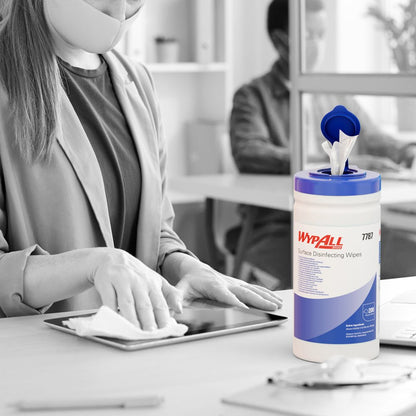 Een gemaskerde persoon desinfecteert met WypAll® oppervlaktedesinfectiedoekjes (dispenseremmer, 200 vellen) van Kimberly-Clark GmbH een tablet op een bureau, terwijl de bak zichtbaar is en een andere persoon op de achtergrond aan een computer werkt.