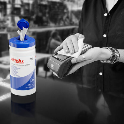 Iemand gebruikt een WypAll® oppervlaktedesinfectiedoekje (Kimberly-Clark GmbH) uit een open dispenserbak om een kaartterminal te desinfecteren. De witte dispenseremmer (200 doekjes) staat op de toonbank, de achtergrond is wazig.