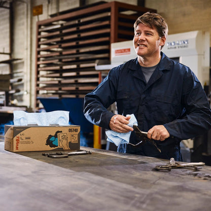 Een man in een donkere overall staat bij een werkbank en reinigt een gereedschap met een blauw WypAll® X60 General Clean doek van Kimberly-Clark GmbH. Een doos met het opschrift "Centrale uitname / Blauw - 1 rol x 150 vellen" staat naast verschillende metalen gereedschappen op de werkbank.