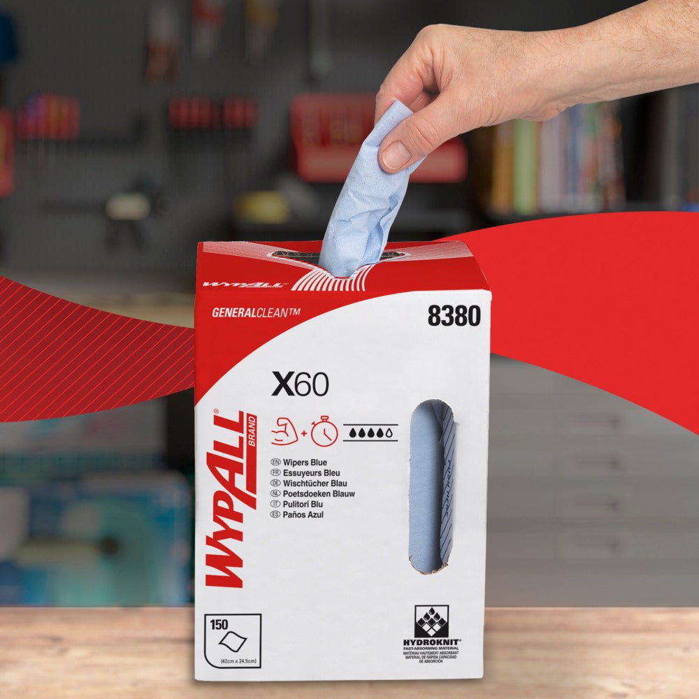 Een hand trekt een blauw WypAll® X60 General Clean vel uit een wit-rode dispenserdoos van Kimberly-Clark GmbH met het opschrift "8380", "150 vellen" en "Centrale uitname". Op de achtergrond is een onscherpe werkplaats met planken te zien.