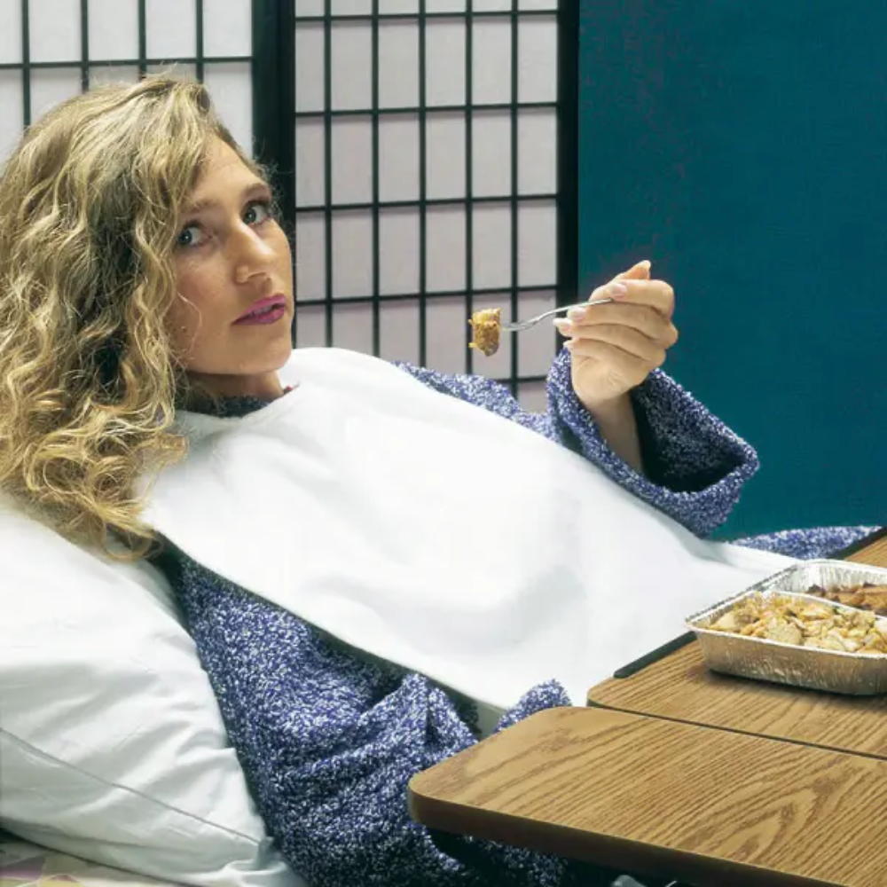 Een persoon met krullend haar ligt in bed tegen een kussen geleund, draagt een blauwe trui en een Servocare kledingbeschermer voor volwassenen van Servoprax GmbH. Ze eet met een vork voedsel uit een foliebakje. Voor hen staat een houten tafel en op de achtergrond een Shoji-scheidingswand.