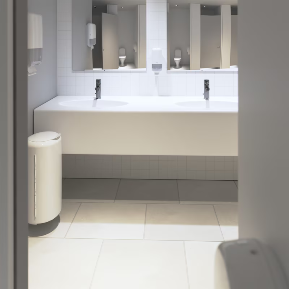 Het openbare toilet presenteert zich in een modern Elevation-ontwerp met wit betegelde muren en vloeren. Een lange witte werkblad herbergt twee wastafels onder een grote spiegel, evenals een Tork 553000 dispenser voor zigzag-handdoeken Elevation H3 van TORK – Essity Professional Hygiene Germany GmbH, die een efficiënte enkelveluitgifte voor papierhanddoeken biedt. Een praktisch geplaatste afvalbak bevindt zich nabij de ingang.