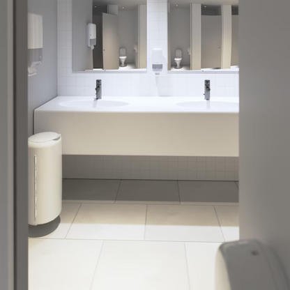 Het openbare toilet presenteert zich in een modern Elevation-ontwerp met wit betegelde muren en vloeren. Een lange witte werkblad herbergt twee wastafels onder een grote spiegel, evenals een Tork 553000 dispenser voor zigzag-handdoeken Elevation H3 van TORK – Essity Professional Hygiene Germany GmbH, die een efficiënte enkelveluitgifte voor papierhanddoeken biedt. Een praktisch geplaatste afvalbak bevindt zich nabij de ingang.