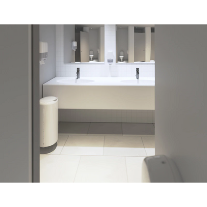 Een schone, moderne openbare toilet beschikt over een lange witte wastafel met twee wasbakken. Boven de wasbakken zijn grote spiegels geplaatst. Aan de muur zijn Tork 553100 Mini-dispensers voor zigzag-handdoeken Elevation H3 van Essity Professional Hygiene Germany GmbH bevestigd, en links daarvan staat een prullenbak. De vloer is betegeld met lichte tegels en heeft een reliëfontwerp.