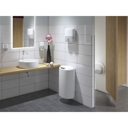 Eine moderne öffentliche Toilette mit weiß gefliesten Wänden und dunkelgrauem Boden ist ein Highlight des Elevation-Designs. Sie verfügt über eine Holzablage mit rundem Waschbecken, großem Spiegel, Tork 555000 Spender für Mini-Jumbo-Toilettenpapier Elevation T2 von Essity Professional Hygiene Germany GmbH, sowie einen Seifenspender, Handtuchspender, Blumenvase, Abfalleimer und eine teilweise geöffnete Kabinentür.
