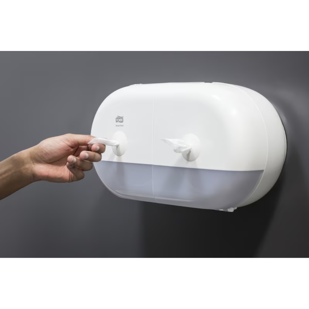 Een persoon trekt een papieren handdoek uit een elegante witte Tork SmartOne® 682000 Mini Dubbelrolhouder voor toiletpapier, die over twee vakken beschikt en aan een gladde, donkergrijze muur is bevestigd.