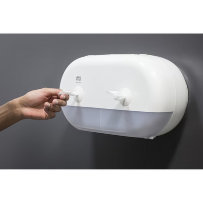 Een persoon trekt een papieren handdoek uit een elegante witte Tork SmartOne® 682000 Mini Dubbelrolhouder voor toiletpapier, die over twee vakken beschikt en aan een gladde, donkergrijze muur is bevestigd.