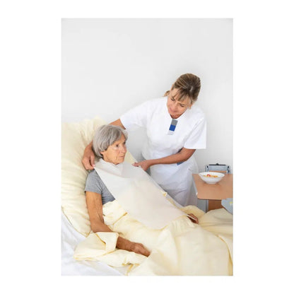 Een verpleegster legt de deken recht voor een oudere vrouw die rechtop in een ziekenhuisbed zit. Beiden lijken aandachtig en geconcentreerd. Een dienblad met een kom en Vala® Fit Tape eenmalige kledingbescherming is te zien.