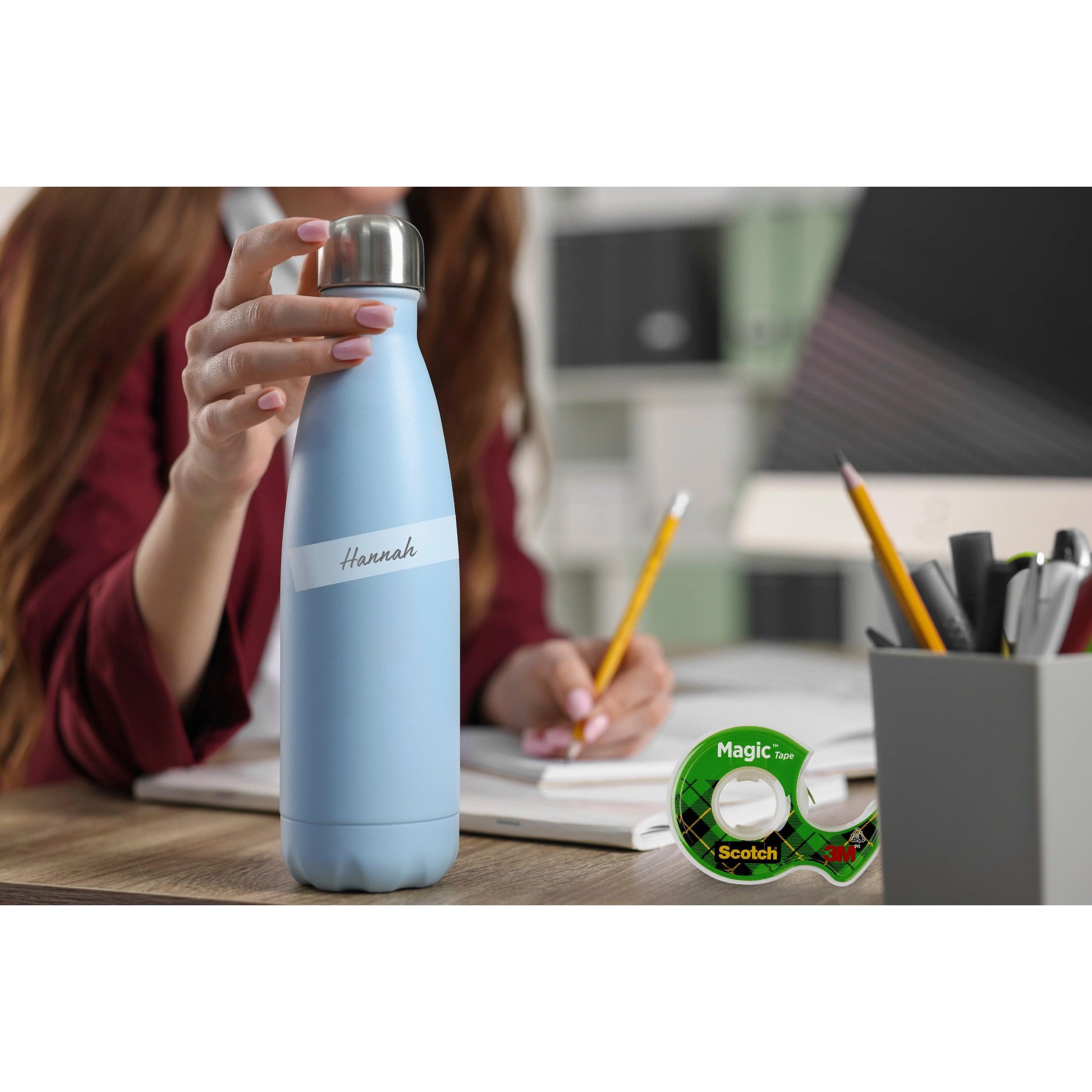 Eine Person hält eine hellblaue isolierte Wasserflasche mit der Aufschrift "Hannah" an einem Schreibtisch mit einem Stifthalter, einem Notizblock und einer Rolle Scotch® Magic™ Unsichtbares Klebeband (19 mm x 7,5 m + Handabroller) von 3M Deutschland GmbH.