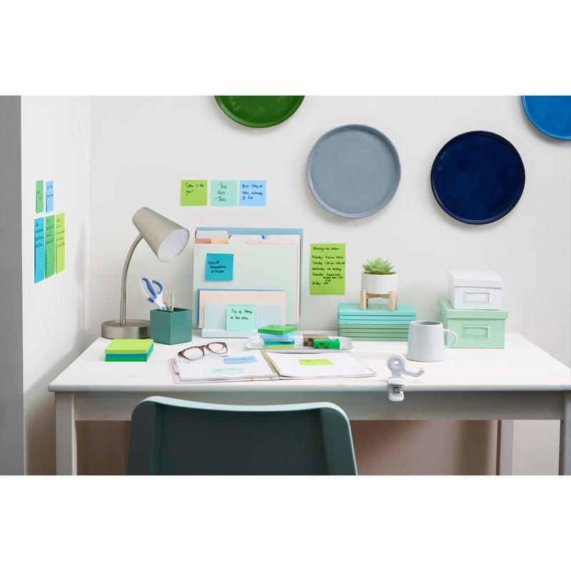 Een opgeruimd wit bureau met kantoorbenodigdheden, lamp, ordners, plant, glazen, dozen en 3M Post-it® Super Sticky Z-Notes (Boost Collection). Kleurrijke borden hangen erboven en Post-it® briefjes zijn aan de muur en op het bureau gerangschikt.