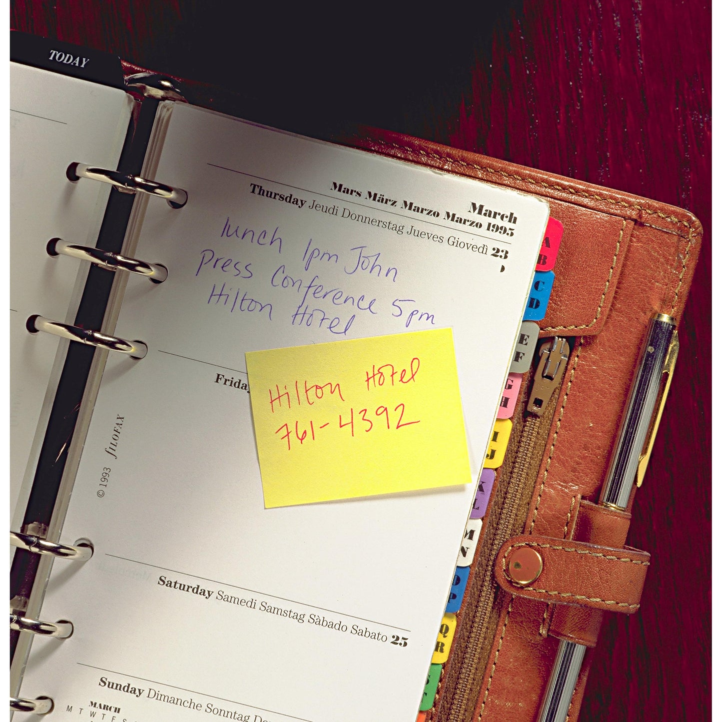 Een geopende leren planner toont handgeschreven donderdagnotities: "Lunch 13 uur John", "Persconferentie 17 uur Hilton Hotel". Op een gele Post-it® Recycling Notes Blok van 3M Deutschland GmbH staat: "Hilton Hotel 761-4392". De scheidingsbladen zijn zichtbaar.