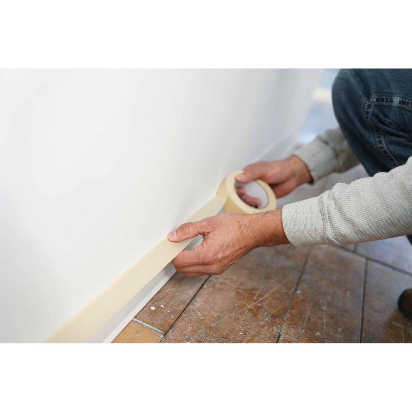 Een persoon plakt Scotch® Afplaktape Classic (48 mm x 50 m, 70% PEFC) van 3M Deutschland GmbH langs de plint van een witte muur naast een tegelvloer en bereidt het oppervlak zorgvuldig voor op het schilderen.