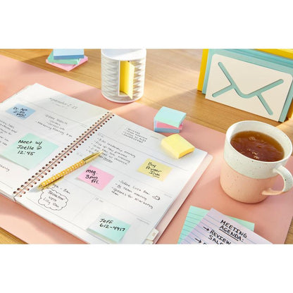 Een open planner met handgeschreven notities, een pen, 3M Deutschland GmbH Post-it® Recycling Notes (76 mm x 127 mm, 100% PEFC), een kopje thee, een kaartje met de tekst "MEETING AGENDA" en bureauaccessoires op een lichtroze bureau-onderlegger.