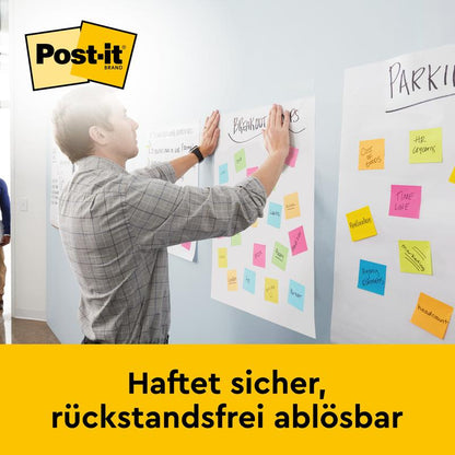 Een man drukt een vel papier met kleurrijke Post-it-notities tegen een muur en presenteert daarbij het 3M Deutschland GmbH Post-it® Super Sticky zelfklevende vergaderblok/flipover voor wandmontage 566 (58,4 x 50,8 cm, 20 vellen, 2 blokken, incl. Command™-strips).