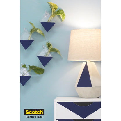 Een lamp met geometrische voet staat op een witte commode met blauwe driehoeken; vier aan de muur bevestigde driehoekige plantenbakken met groene planten hangen aan een lichtblauwe muur. Het logo van 3M Scotch® afplaktape voor gevoelige oppervlakken verschijnt in de linker onderhoek.