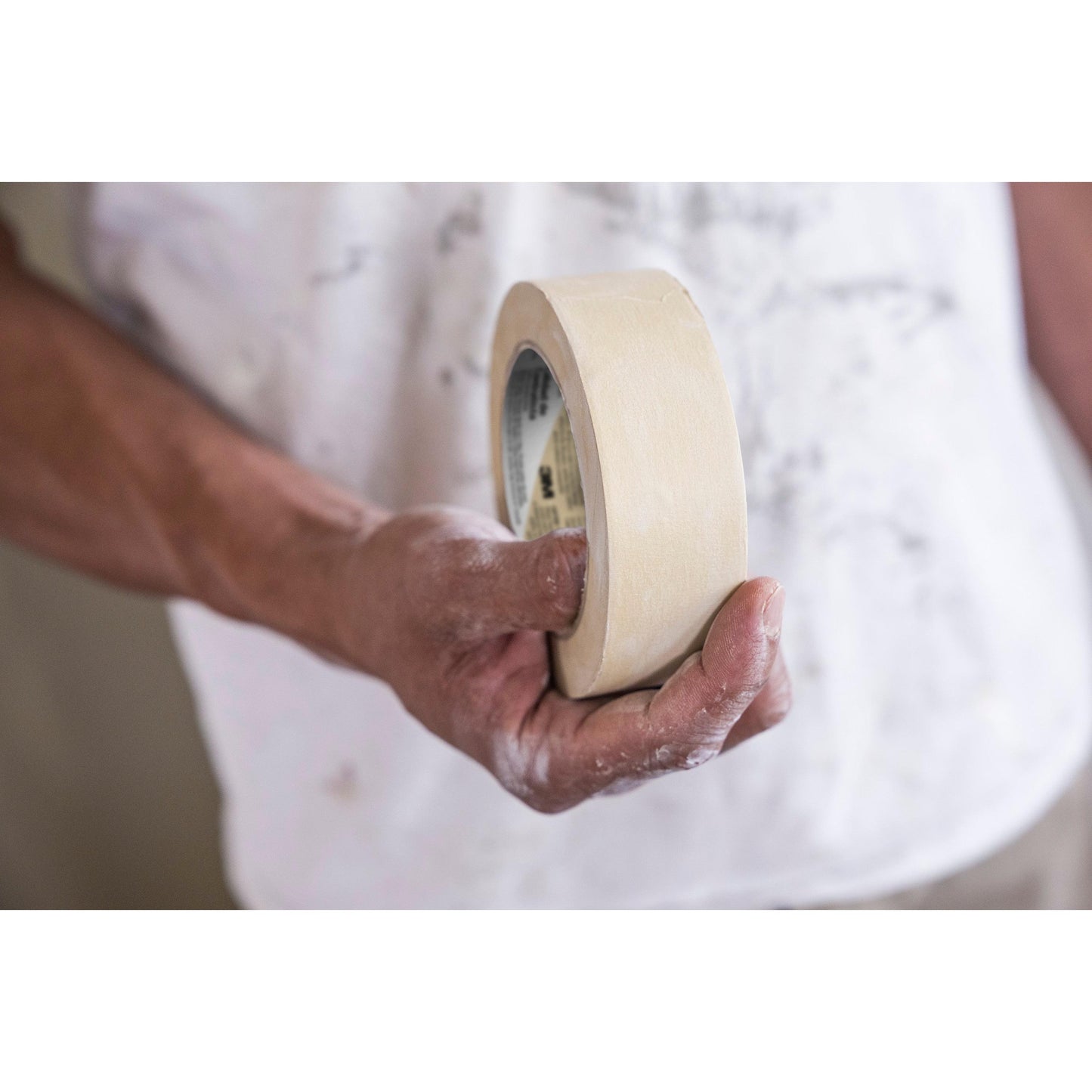 Een persoon met met verf besmeurde handen houdt een rol Scotch® Afplaktape Classic (48 mm x 50 m, 70% PEFC) van 3M Deutschland GmbH vast en bereidt zich voor op schilderwerk in een wit, met verf besmeurd shirt met een wazige achtergrond.
