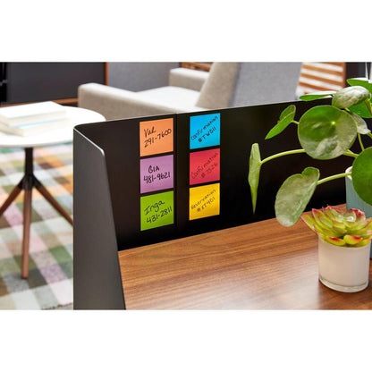 Een zwarte metalen verdeler op een houten tafel toont zes verpakkingen 3M Deutschland GmbH Post-it® Notes, Geel, 76x76mm, 90 vel/blok, PEFC-gecertificeerd. Groene potplanten versieren het bureau, op de achtergrond staan boeken en een stoel.