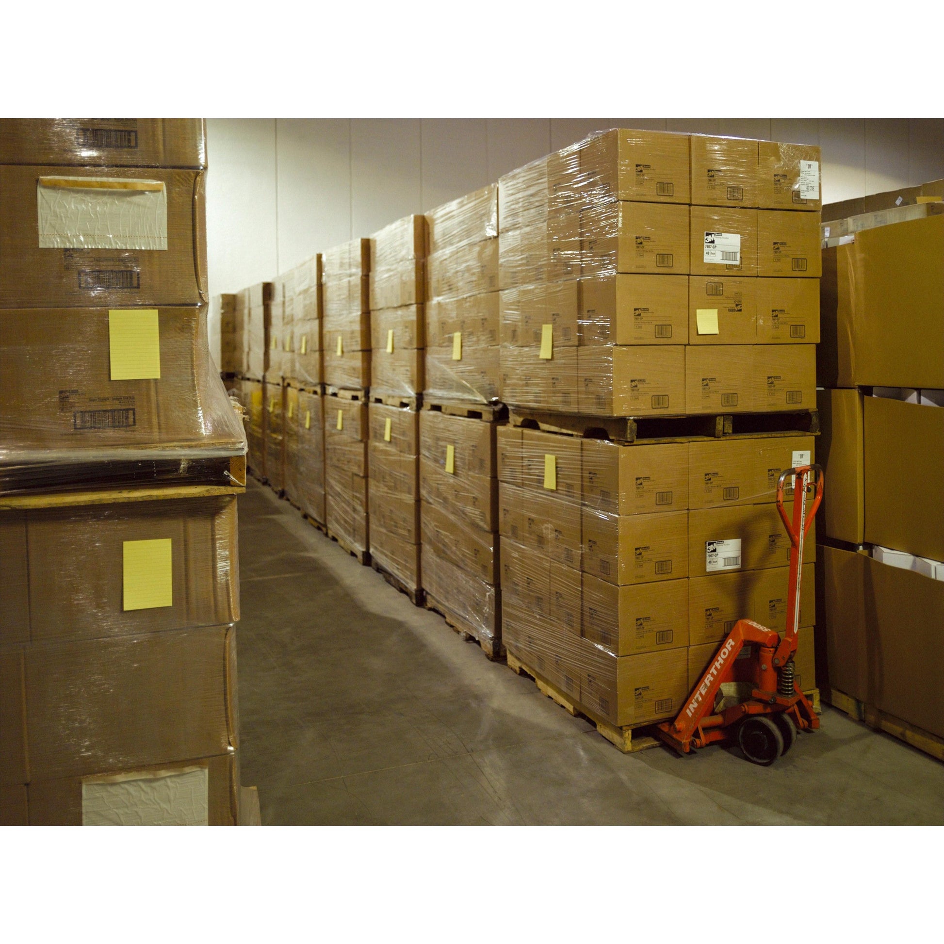 In einem Lagerhaus stapeln sich große Post-it® Super Sticky Notes der 3M Deutschland GmbH in Pappkartons auf Paletten, eingewickelt in Plastik. Ein roter Hubwagen steht in der Nähe und einige Kartons sind mit gelben Etiketten versehen.