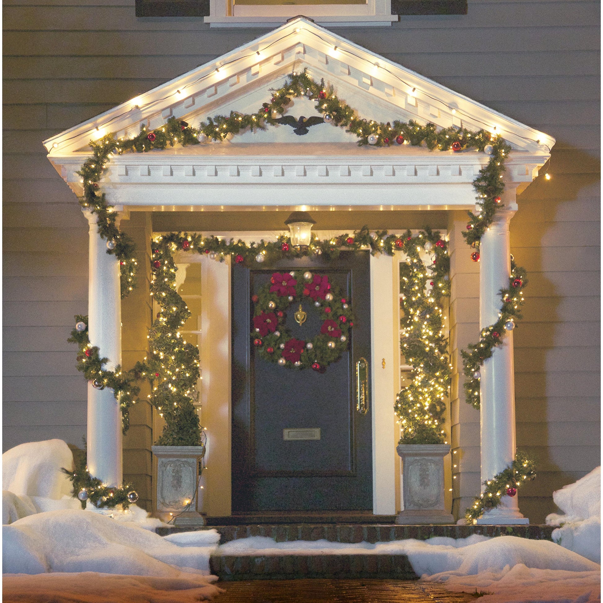 Ein gemütlicher Weihnachtseingang erstrahlt mit Lichterketten, die mit Command™ Outdoor Lichter-Clips, 17017CLR-AWPU (8 Clips + 10 Streifen) von 3M Deutschland GmbH aufgehängt wurden, Schnee auf den Stufen und Girlanden, die die dunkle Tür einrahmen, für eine charmante Außenbeleuchtung Befestigung.