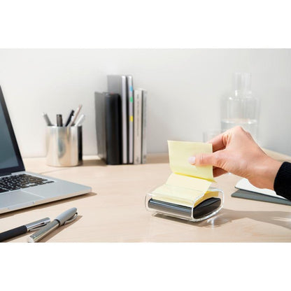 Een hand trekt een gele Post-it® Super Sticky Z-Note (76x76 mm) uit de zwarte PRO Post-it® Z-Notes Dispenser van 3M Nederland B.V. op een opgeruimd bureau met laptop, pennen, boeken, glazen karaf en beker, netjes op de achtergrond.